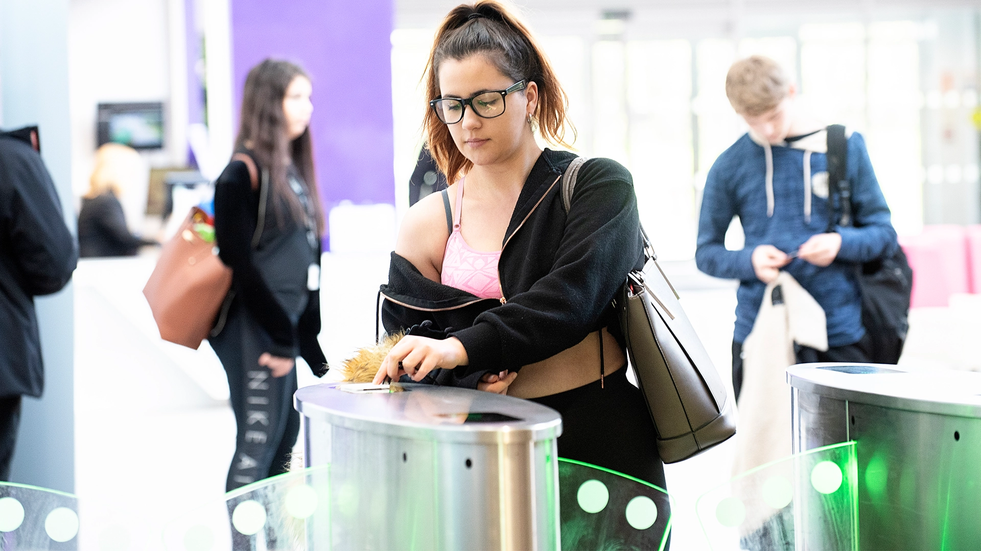 A student taps an ID card on an electronic access gate inside a bright campus entrance area, with other students walking through or waiting nearby.