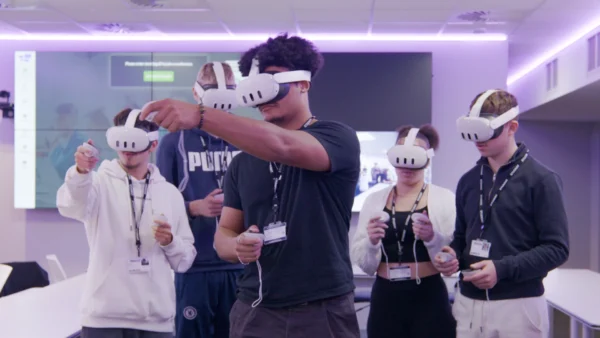 A group of students stand in an immersive classroom wearing virtual reality headsets and holding VR controllers, with large screens and purple ambient lighting in the background.