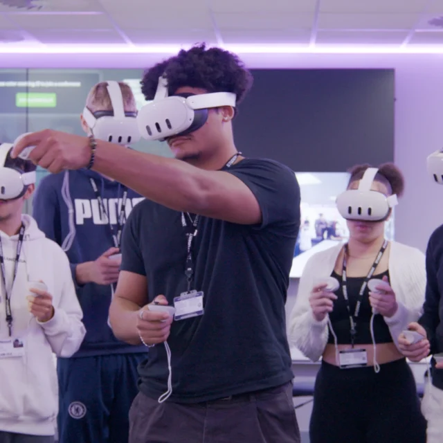 A group of students stand in an immersive classroom wearing virtual reality headsets and holding VR controllers, with large screens and purple ambient lighting in the background.