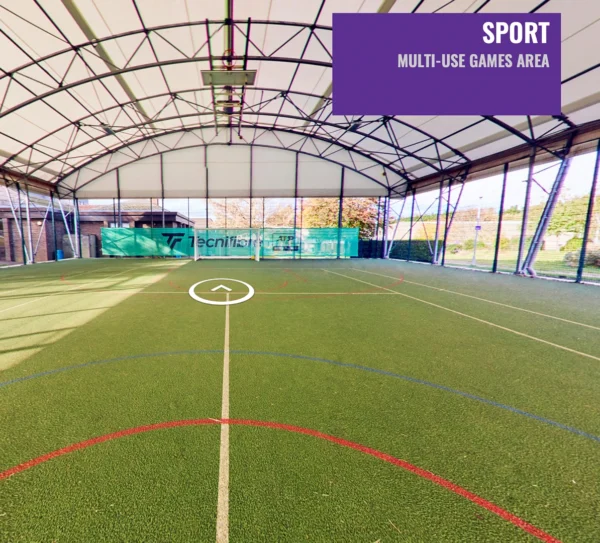 An indoor multi‑use games area with artificial turf marked for sports activities, housed under a high arched roof structure.