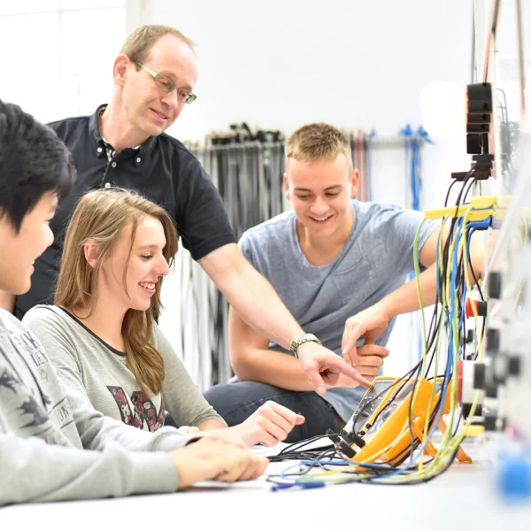 A group of learners sit at a workbench using electronic equipment while an instructor stands beside them and points to the controls.