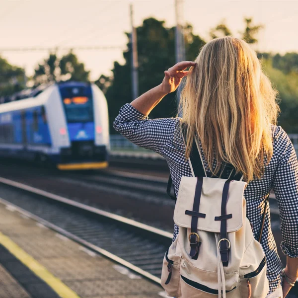 A person with long hair wearing a backpack stands on a train platform, facing an approaching blue and white train.