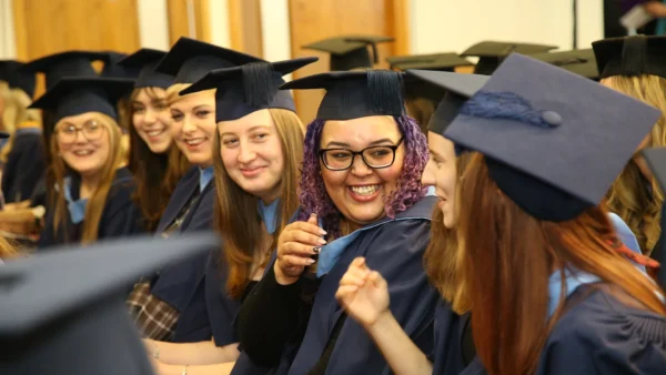 A group of graduates seated in rows wearing caps and gowns, facing forward and interacting with one another during a ceremony.
