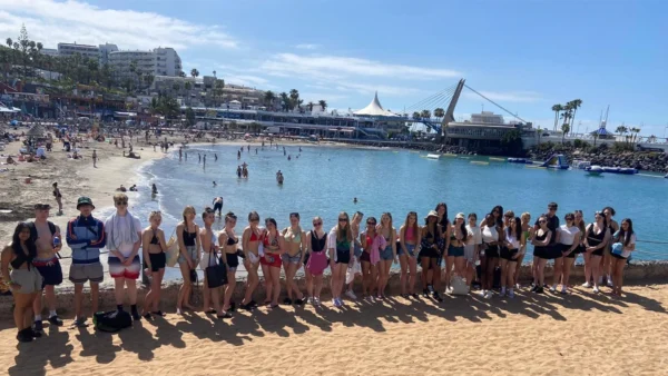 A large group of students standing in a line on a sandy beach with a calm bay and busy waterfront in the background.