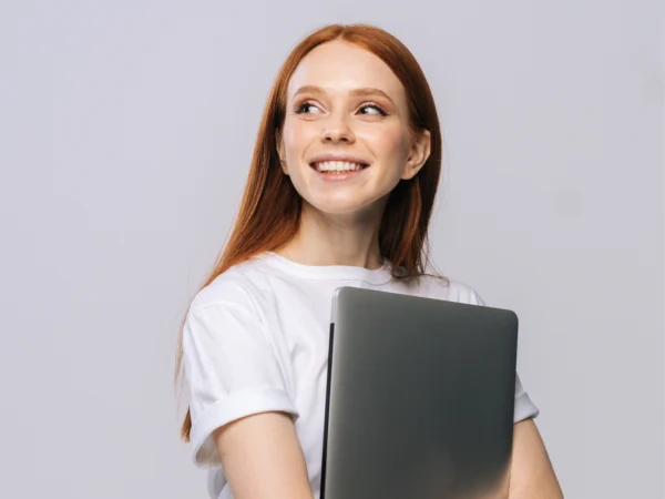 Person wearing a plain white shirt holding a closed laptop against a neutral background.