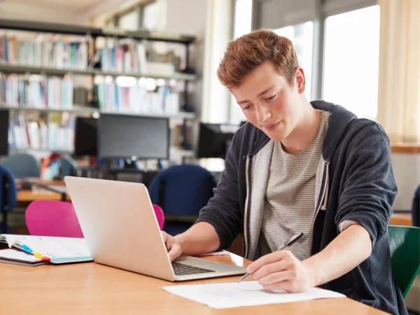 Person working at a desk with a laptop and papers in a library setting.