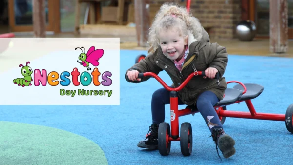 A young child wearing a coat rides a red tricycle on an outdoor play surface. The Nestots Day Nursery logo, featuring a cartoon caterpillar and butterfly, appears on the left side of the image.