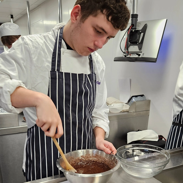Person wearing a chef’s jacket and striped apron, stirring a bowl of chocolate mixture with a wooden spoon in a professional kitchen.