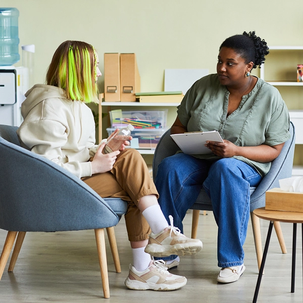 Two people seated in armchairs facing each other, talking indoors, with one holding a clipboard, in a light, informal meeting space.