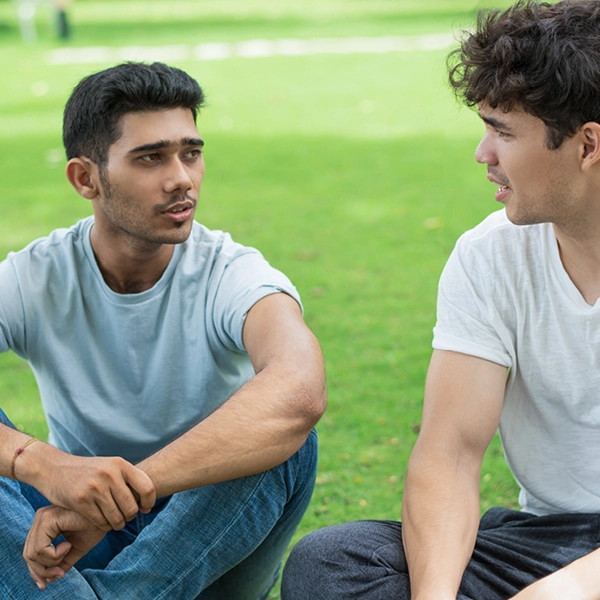 Two people sitting on grass outdoors, facing each other and talking, with a green lawn in the background.