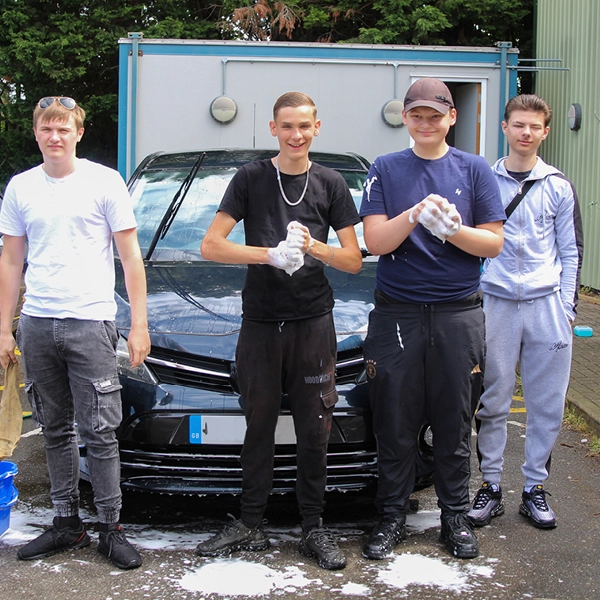Five people standing in front of a dark car being washed outdoors, with soap on the ground and some holding cleaning sponges, in front of a portable building.