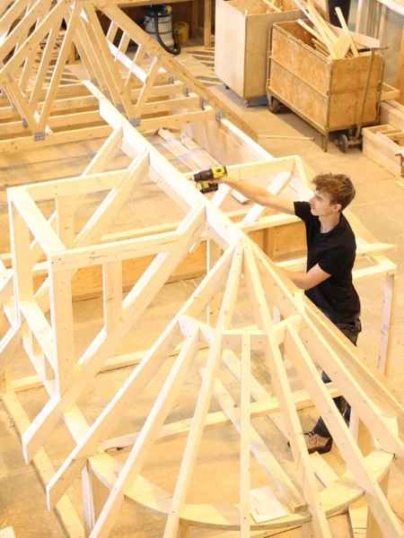 Person using a power drill to assemble a large wooden roof‑frame structure inside a construction workshop, with timber frameworks and tools visible around the space