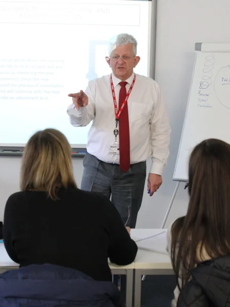 Tutor delivering a teacher education lesson to a small group of mature students in a classroom, using a presentation screen and a flip chart.