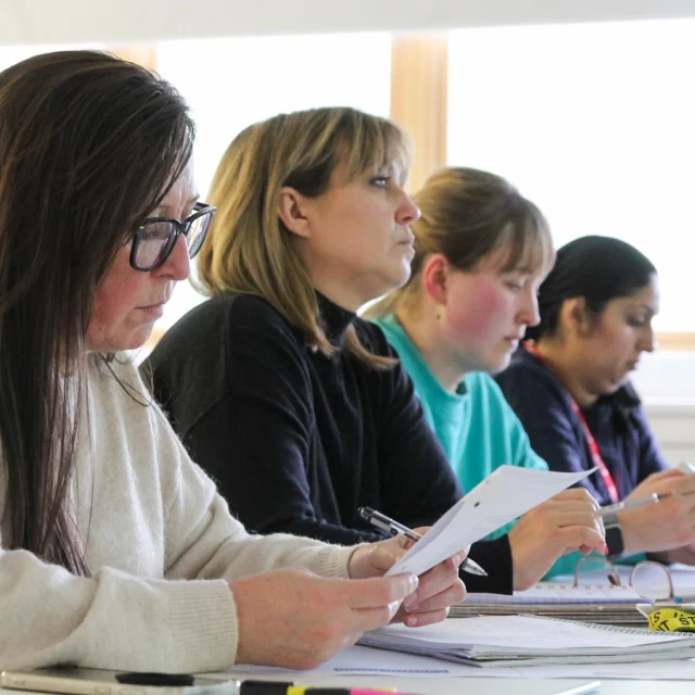 Adult learners seated at desks in a classroom, reading papers and taking notes during a higher education lesson.
