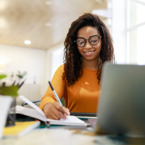 Student writing in a notebook while using a laptop at a desk in a bright study space