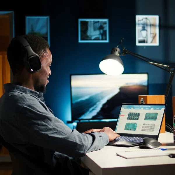 Person wearing headphones using a laptop at a desk in a home workspace, with a desk lamp and external monitor in the background.