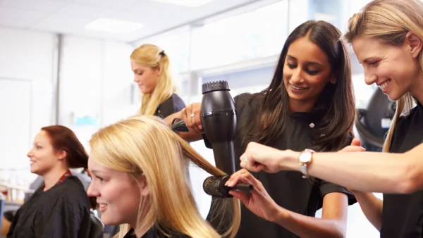 Learners practise hair styling in a training salon, using a hairdryer and brush while others observe nearby.