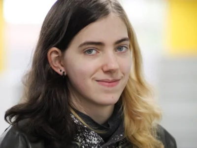 Close-up portrait of a person with shoulder-length two-tone hair, wearing a dark jacket and patterned top, looking towards the camera against a softly blurred indoor background.