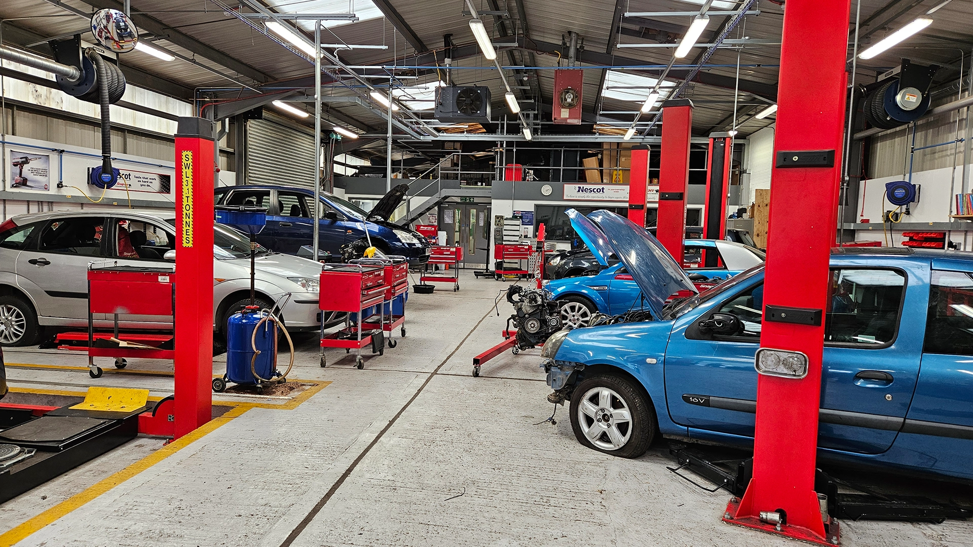 Automotive workshop with cars on lifts, exposed engine components and tool stations throughout the workspace.