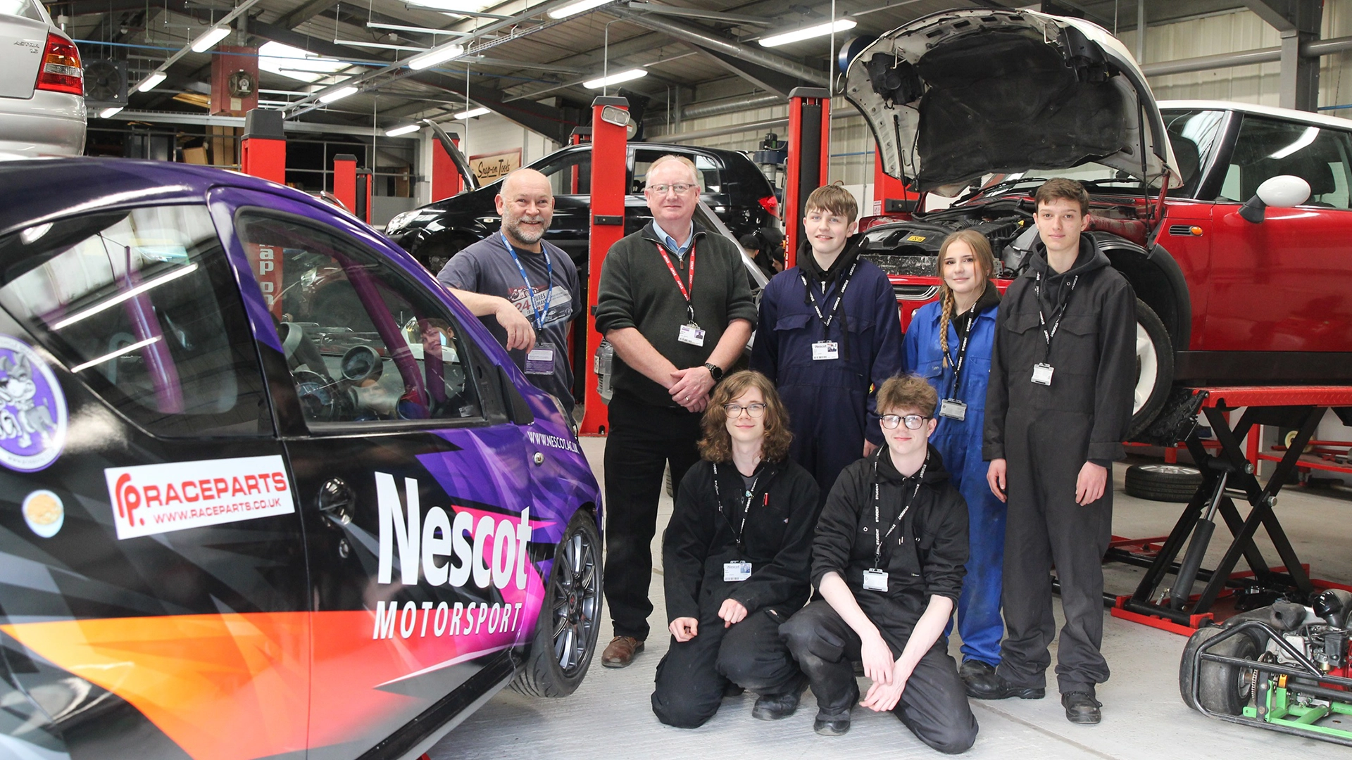 Group of students and staff in a workshop beside a Nescot Motorsport car on a vehicle lift.