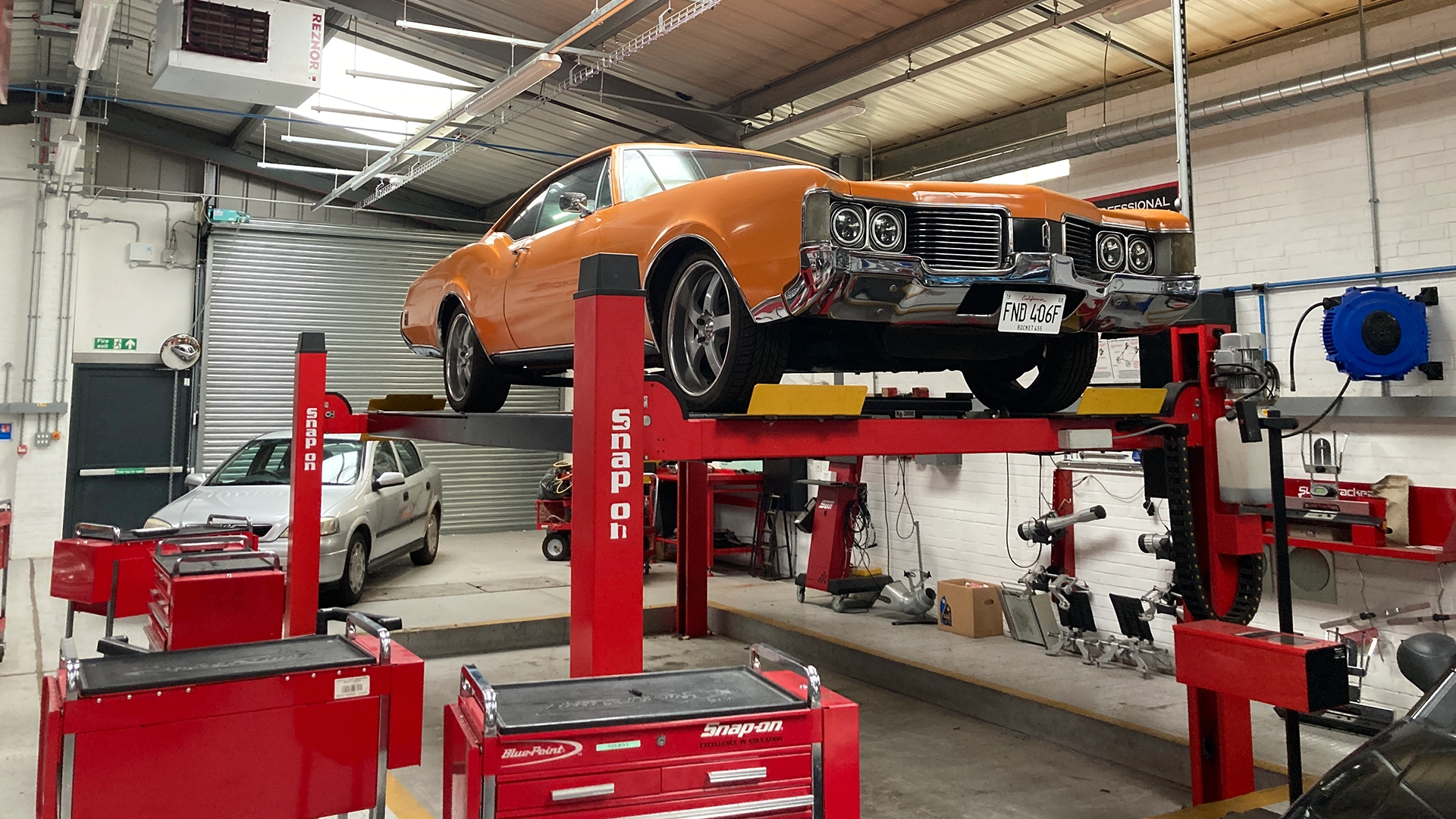 Classic orange car raised on a lift inside an automotive workshop with tools and equipment nearby.