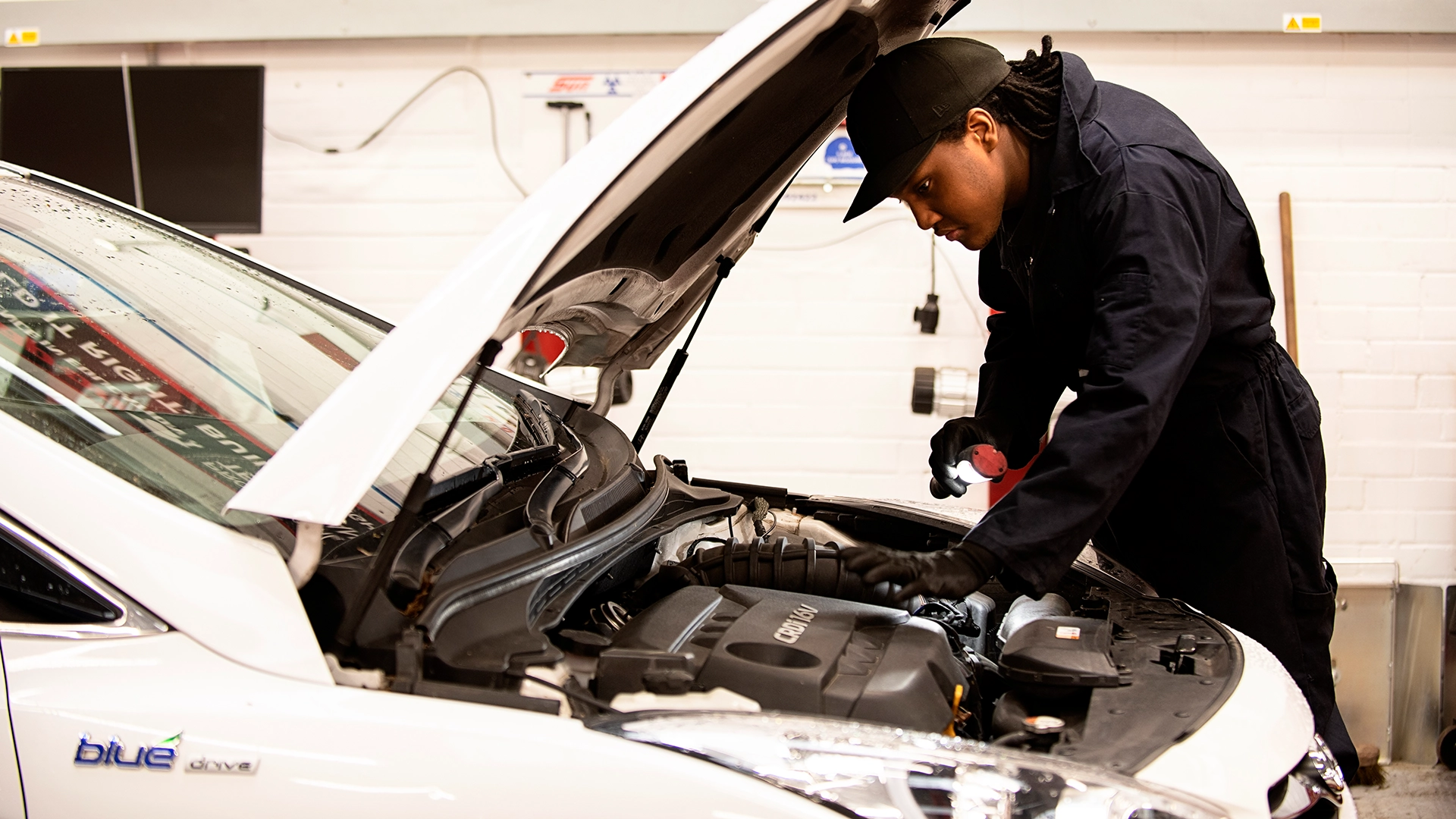 Person inspecting an open car engine using a handheld light in a workshop setting.