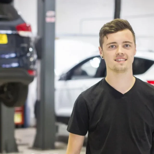 Person standing in a vehicle workshop with cars on lifts in the background, wearing a black T-shirt.