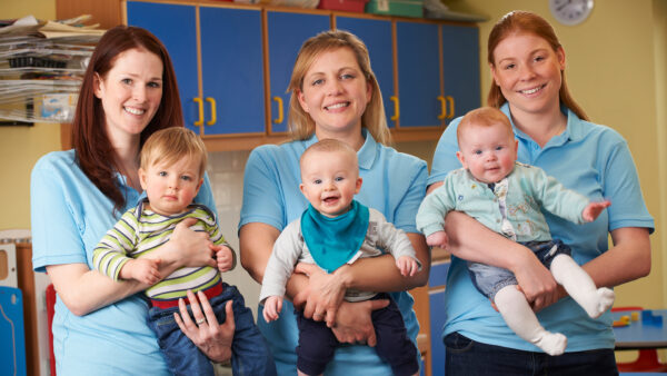 Three adults in light blue uniforms each holding a baby in a nursery classroom, with shelves and equipment in the background.