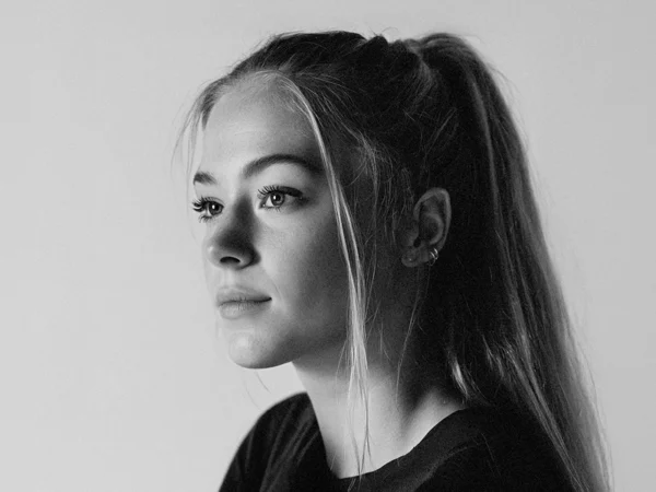 Black-and-white close-up portrait of a person in side profile, facing left, with long hair tied back in a ponytail, wearing small earrings and a dark top, against a plain light background.