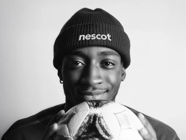 A person wearing a beanie with the word ‘nescot’ on it holds a football close to the camera while standing against a plain background.