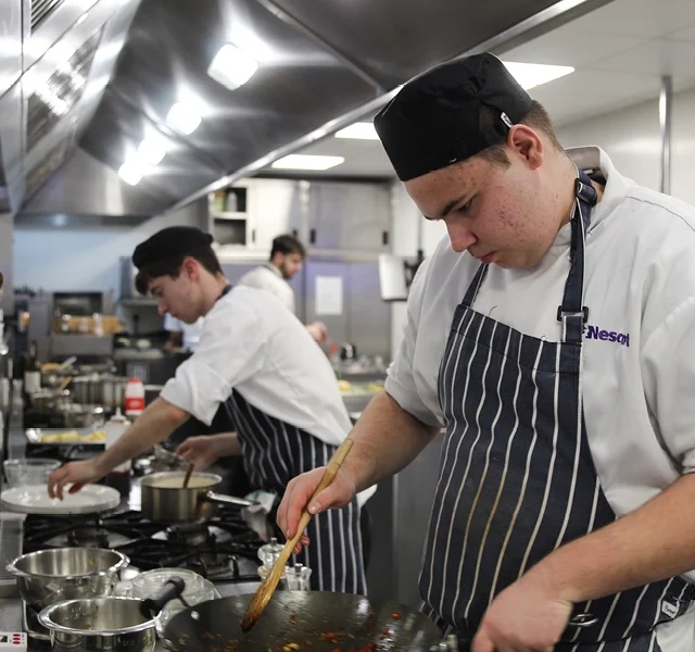 Catering students working in a professional kitchen, preparing food at a stovetop with pans and utensils while wearing chef whites and striped aprons.