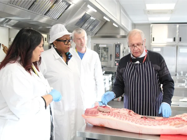 Instructor wearing apron demonstrates meat preparation on a large cut of carcass in a professional kitchen, while three trainees in lab coats and gloves observe; stainless steel equipment and ovens are visible in the background.