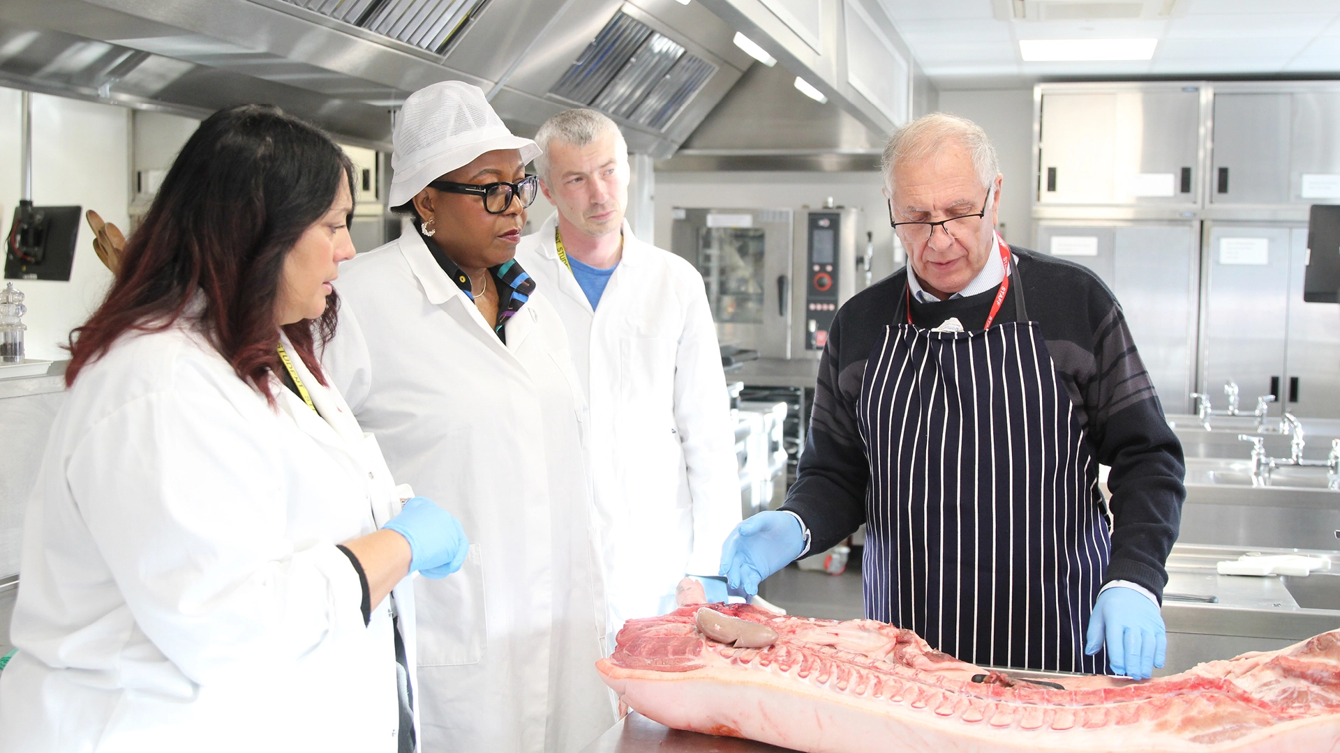 Instructor wearing apron demonstrates meat preparation on a large cut of carcass in a professional kitchen, while three trainees in lab coats and gloves observe; stainless steel equipment and ovens are visible in the background.