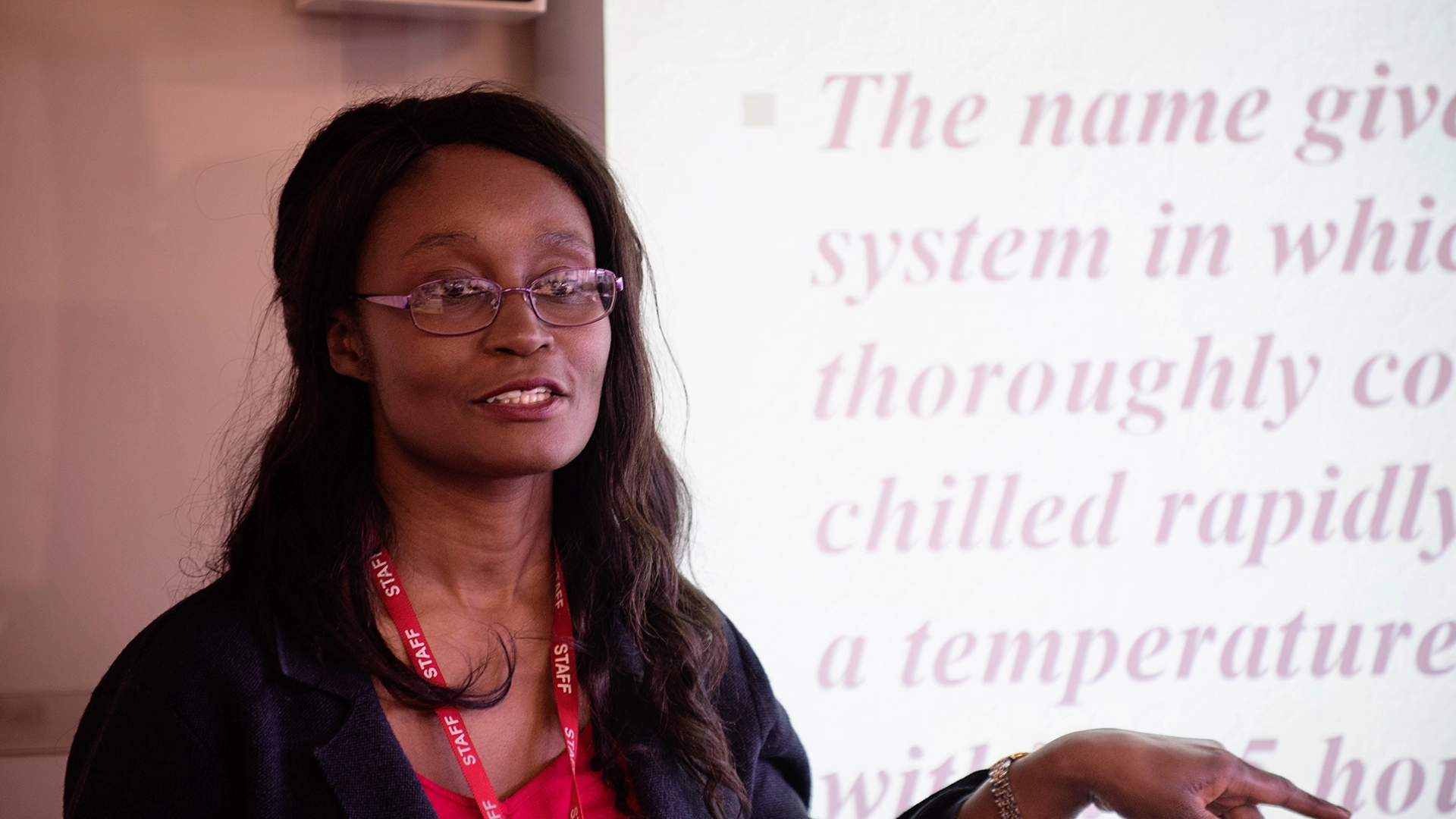 Tutor wearing a staff lanyard stands in front of an interactive whiteboard, pointing while delivering a presentation; blurred text about food safety is visible on the screen behind.