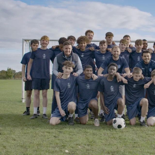 Group of students and staff posing for a team photo on a grass football pitch, wearing matching sports kit, with a goalpost and cloudy sky in the background.