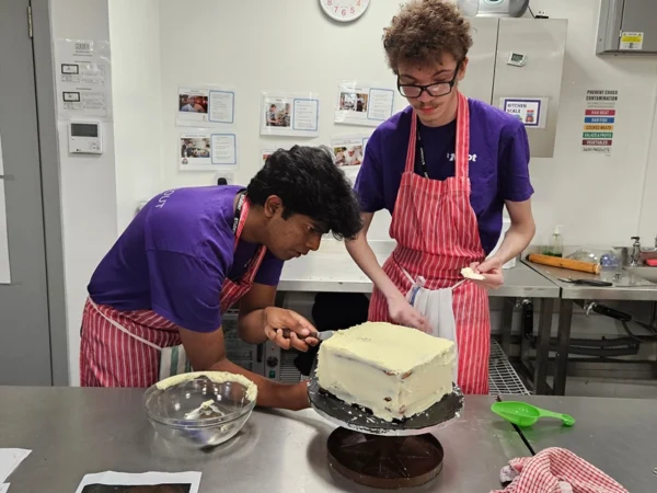 Two people in matching purple tops and red striped aprons ice a square cake on a stand in a commercial kitchen. One smooths frosting on the side with a spatula while the other holds a utensil over the cake. A mixing bowl, green measuring scoop, and utensils sit on the stainless steel work surface. Posters and a wall clock are visible in the background.