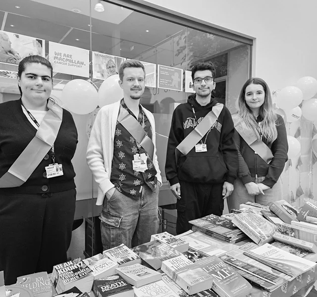Students stand behind a table covered with books and leaflets at a fundraising stall indoors, wearing sashes and ID badges.