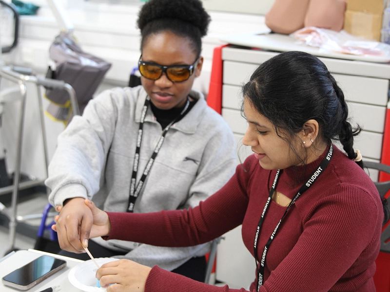 Student practical activity in a classroom, with two people wearing lanyards using a small tool and material at a desk, with equipment and medical supplies in the background.