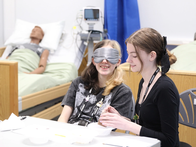 Two students practising a sensory activity in a training room, with one wearing an eye mask while handling an object with guidance from another, with a hospital bed, mannequin, and monitoring equipment visible in the background.