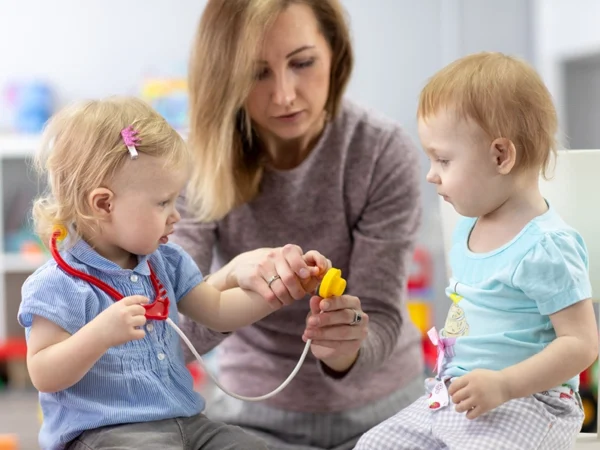 Adult sits with two toddlers during a play activity; one toddler holds a toy stethoscope while the adult helps guide the other toddler’s hand on a small object, with colourful play items in a softly blurred indoor setting.