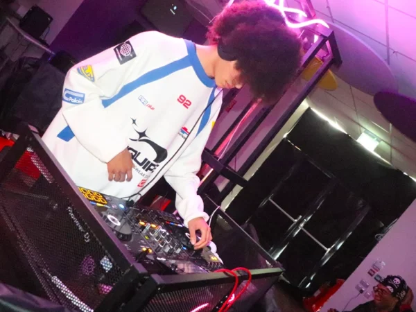 Person using a DJ controller at a booth with pink neon lighting and cables visible.