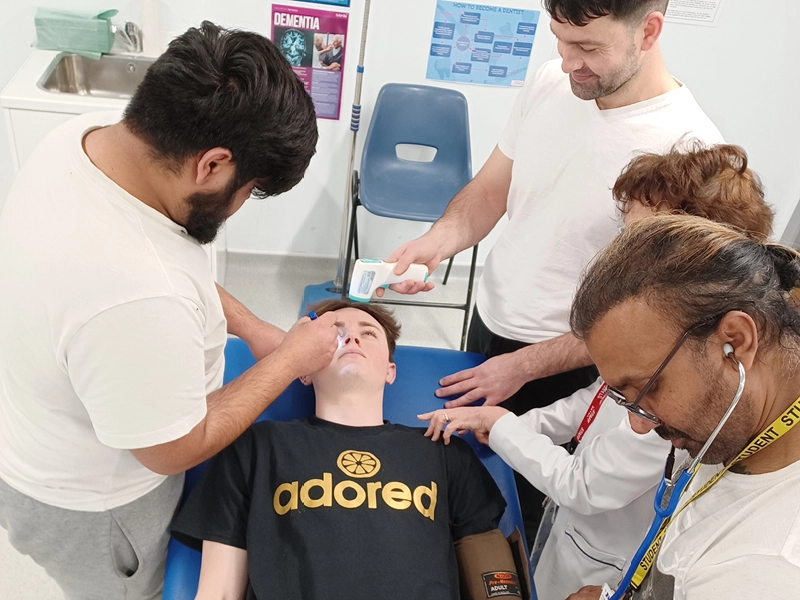 Person lying on an examination couch while several others perform a clinical check in a medical training room; one uses an instrument near the eye, another holds a handheld device, and a third uses a stethoscope and blood pressure cuff. Posters are visible on the wall behind.