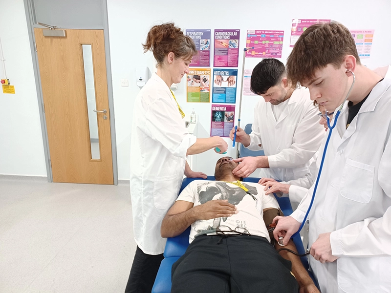 Group of people in a clinical training room examining a person lying on a treatment couch; one checks the eyes with a tool, another uses a stethoscope and blood pressure cuff, while posters about health topics are displayed on the wall behind.