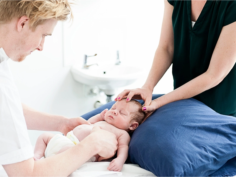 Clinician supports an infant lying on a treatment couch, with hands placed gently on the head and torso, while another clinician observes nearby; a sink and plain clinical setting are visible in the background.