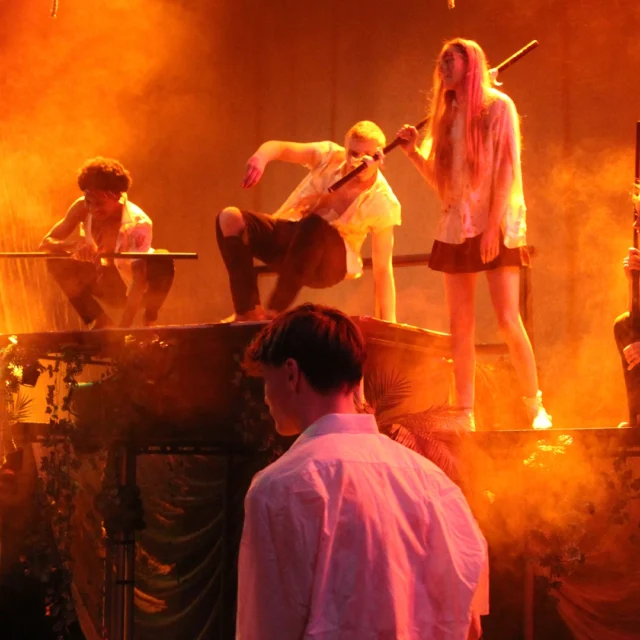 Group of performers stand and crouch on a raised stage set, holding poles, surrounded by theatrical smoke and warm orange lighting; foliage and set pieces suggest an outdoor or jungle scene, with other performers visible in the foreground and background.