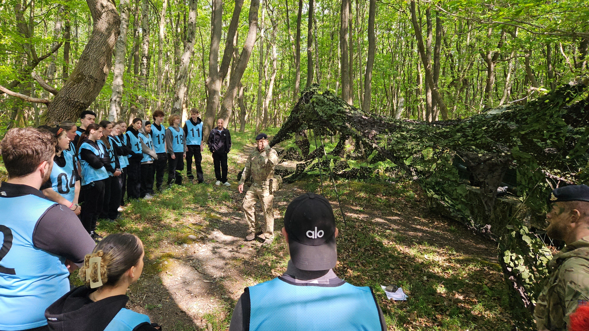 Group of students wearing numbered blue bibs standing in a wooded area, listening to an instructor in camouflage clothing giving instructions beside a camouflaged shelter among trees.