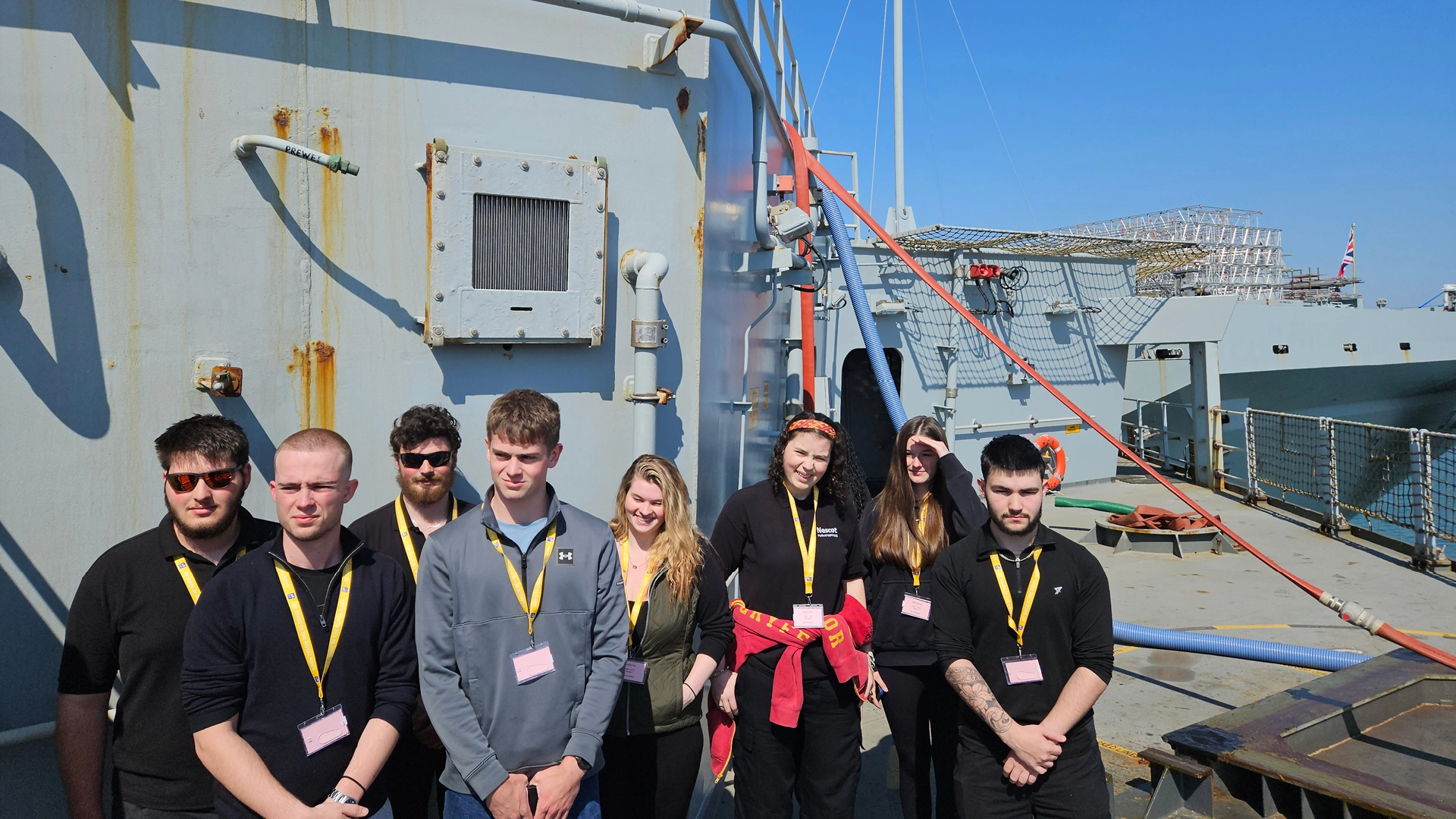 Group of students standing on the deck of a large military vessel, wearing ID lanyards, with industrial equipment, pipes and safety netting visible under a clear blue sky.