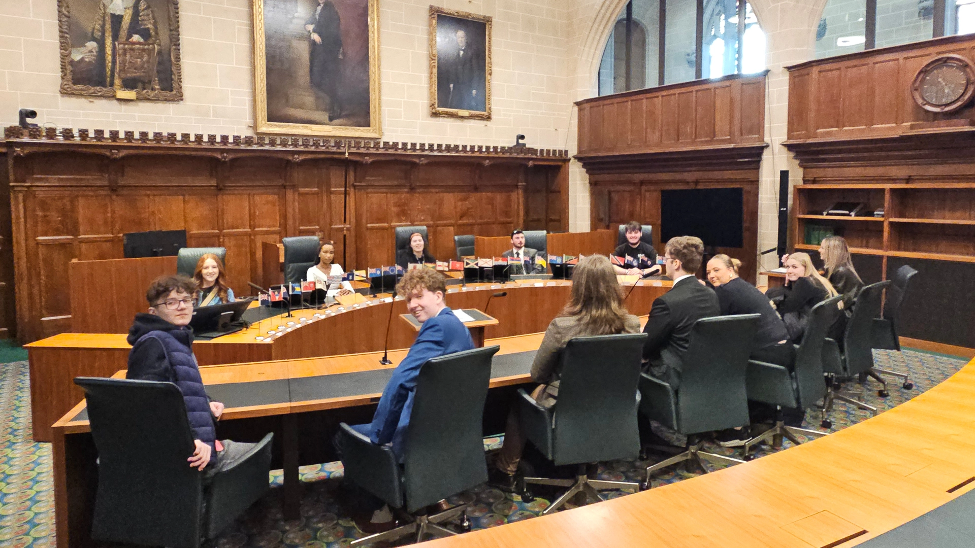 Group of students seated around a large curved desk in a formal wood-panelled legal chamber, with small national flags displayed along the table and framed portraits on the walls.