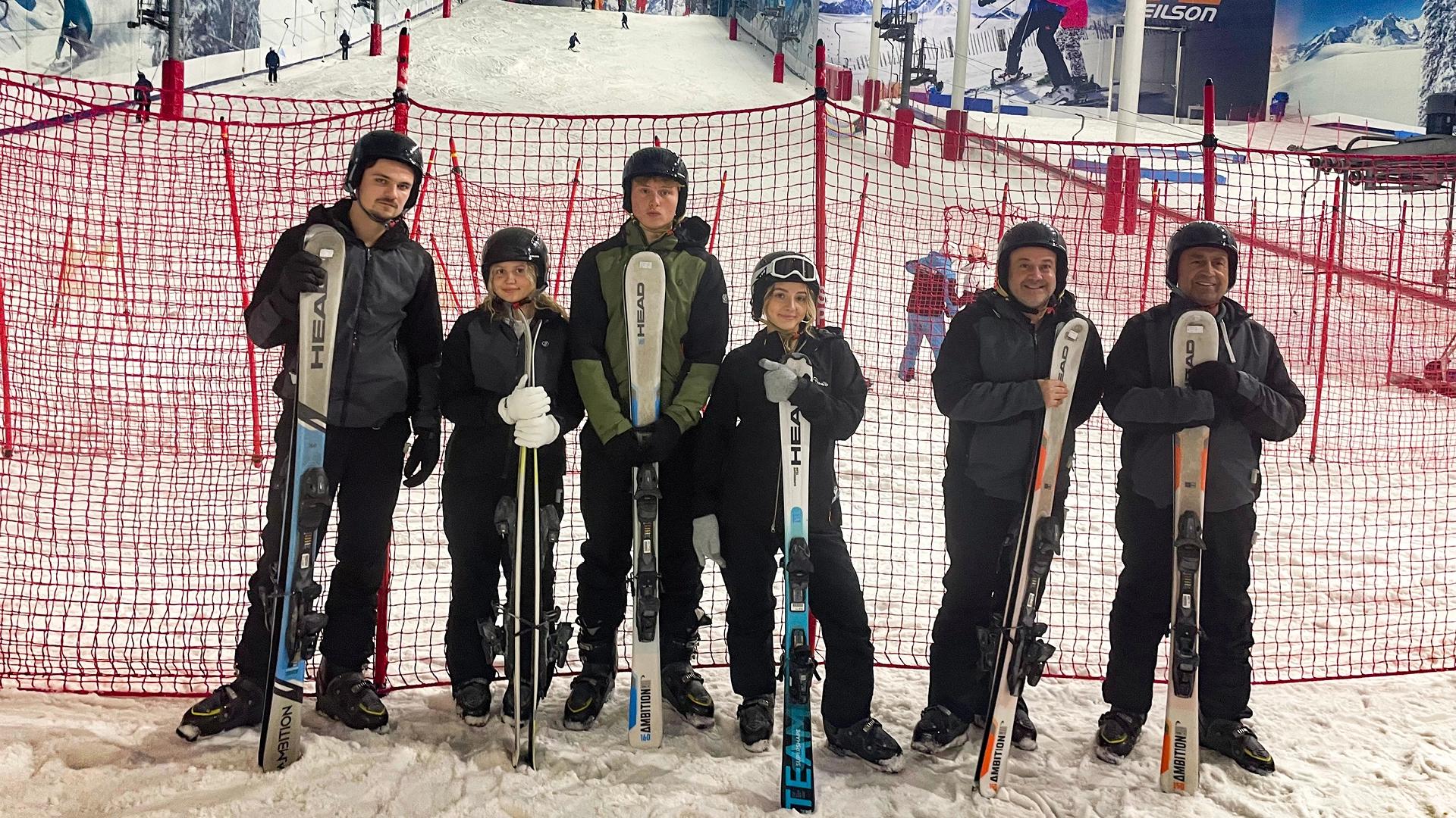 Group of people in winter clothing and helmets standing on an indoor ski slope, holding skis in front of a red safety net, with other skiers and snowy slopes visible in the background.