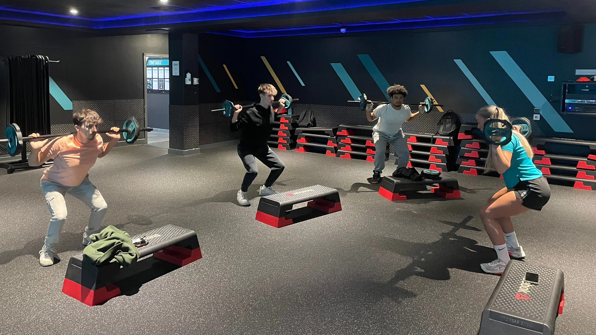 Group of people in a dimly lit fitness studio performing barbell squats on step platforms, with stacked equipment, blue accent lighting, and wall graphics in the background.